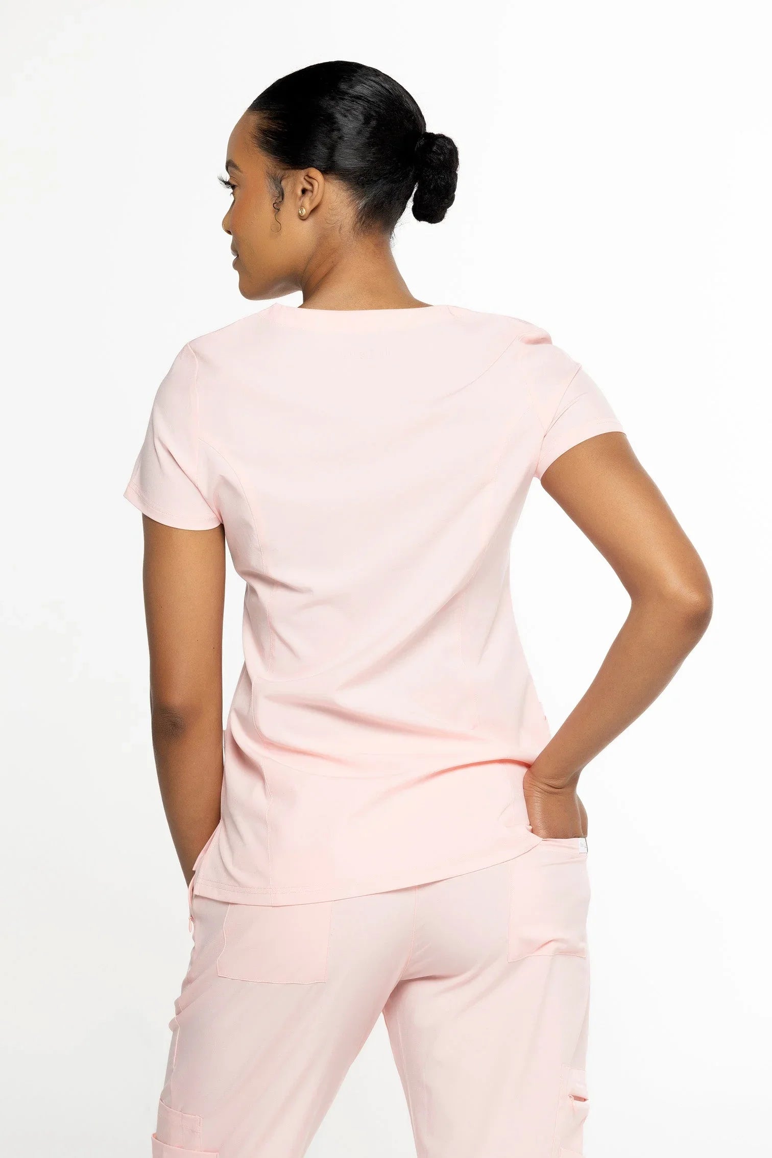 Woman wearing light pink medical scrub top and pants, shown from back with hand in pocket