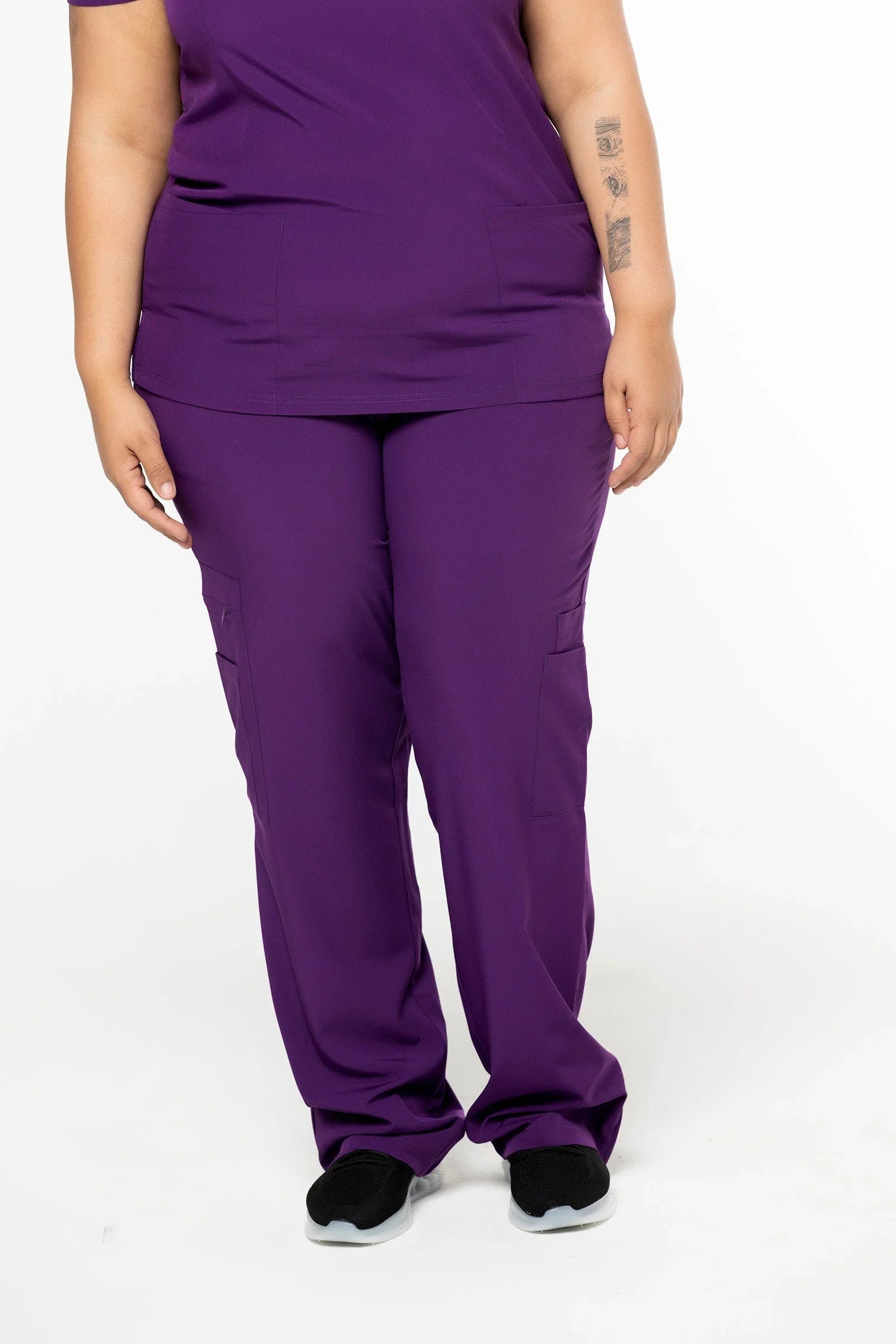 Plus size model wearing purple medical scrub pants and top with black sneakers on white background