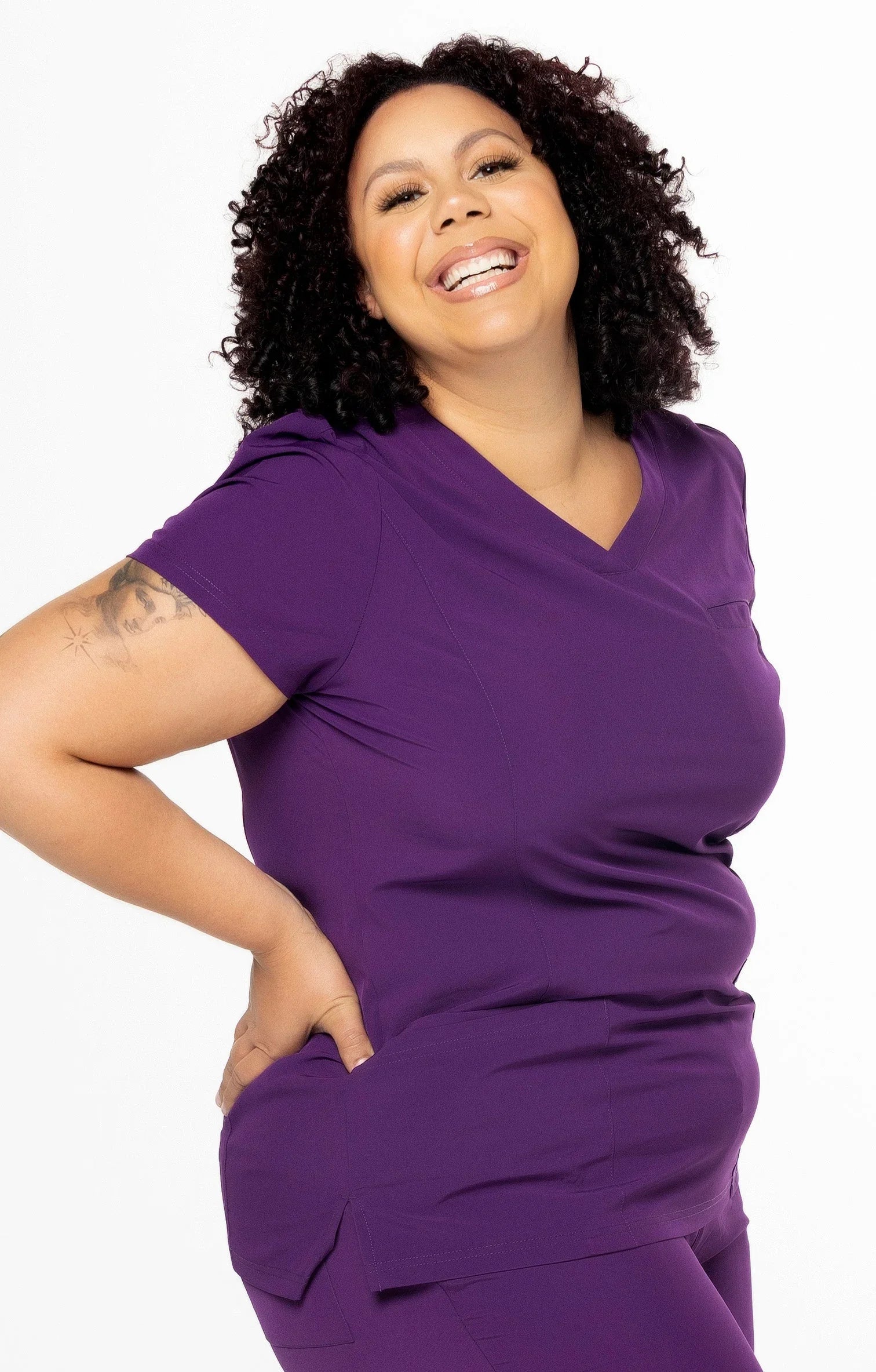 Smiling woman with curly hair wearing purple medical scrubs with tattoo on arm, white background