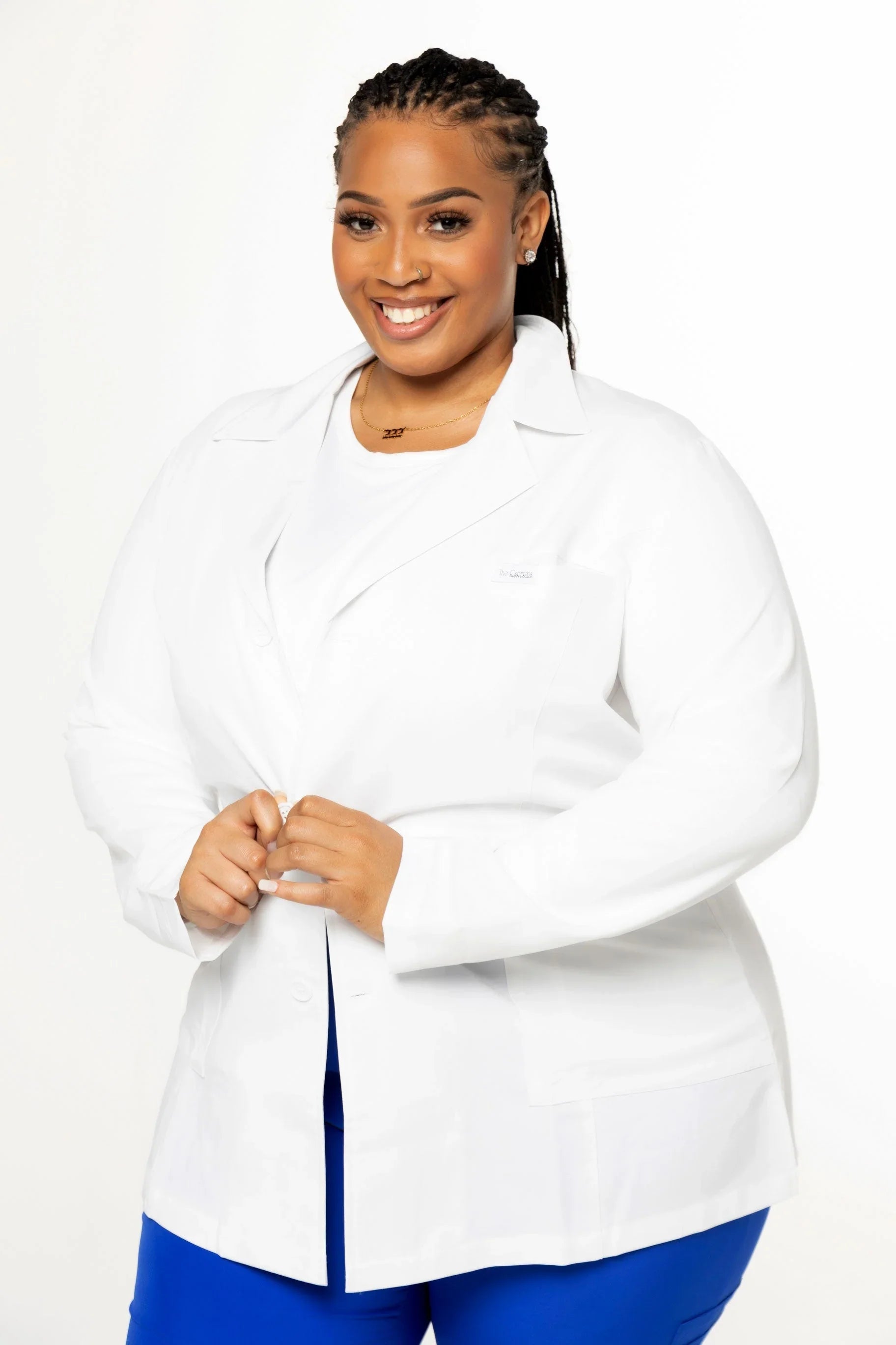 Smiling woman with braided hair wearing a white lab coat and blue pants on white background