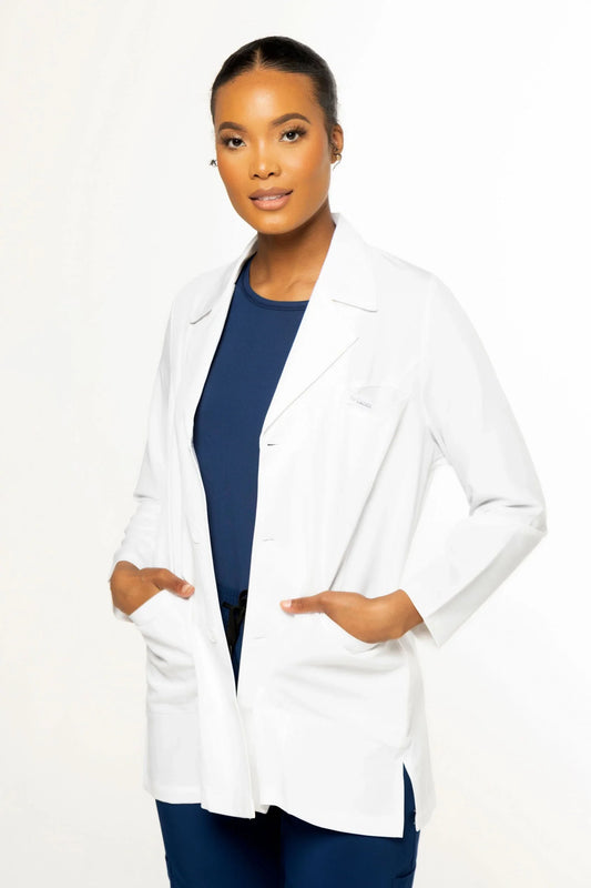 Young woman wearing white medical lab coat over navy blue scrubs posing against white background
