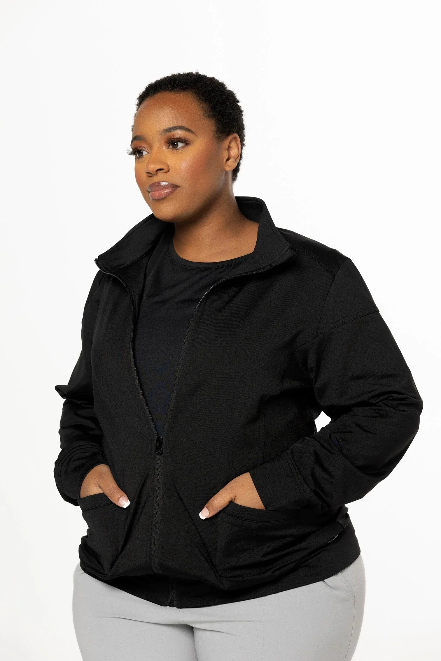 Woman wearing a black zip-up jacket with hands in pockets, posing against white background