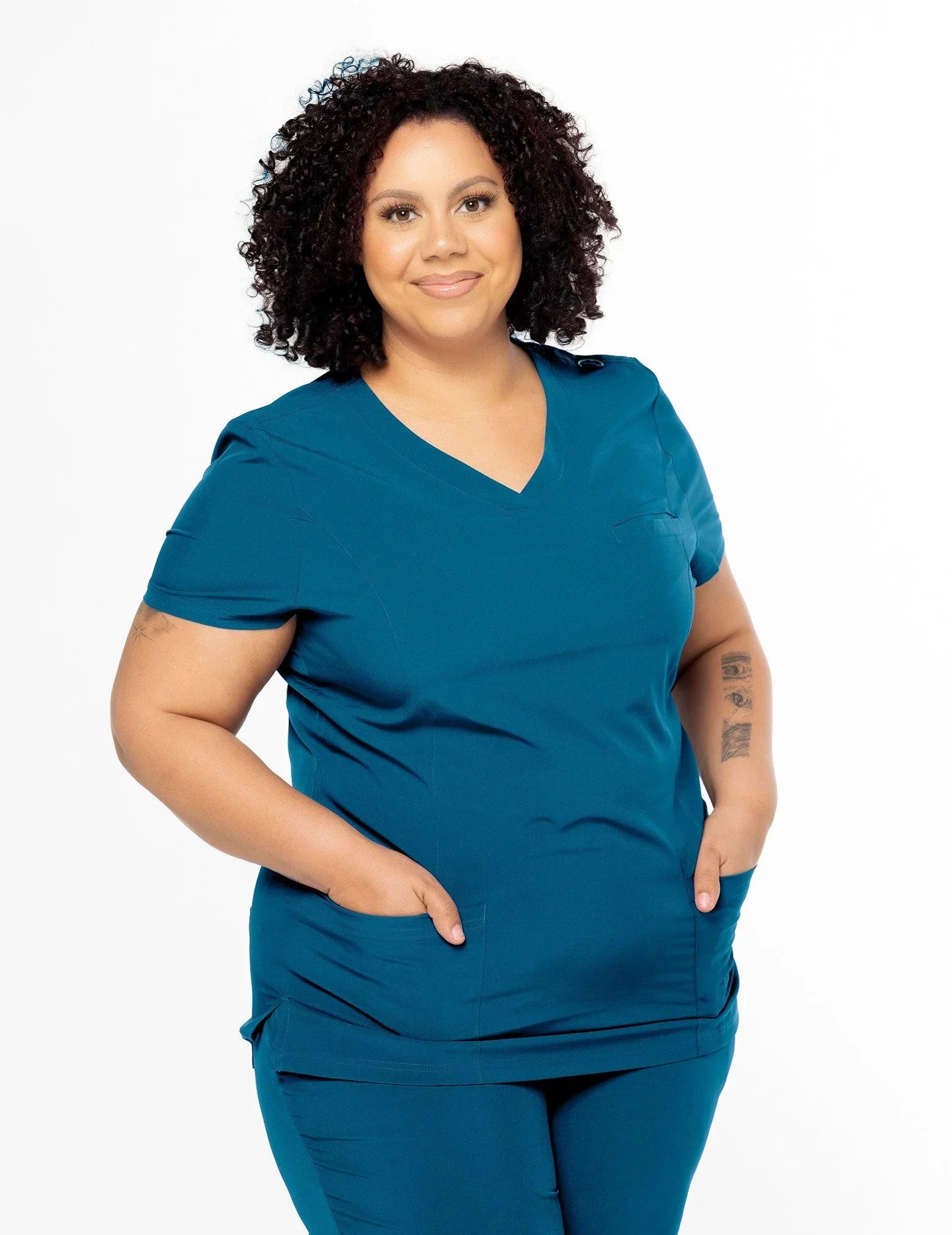 Curly-haired woman wearing teal medical scrubs with pockets, standing against a white background