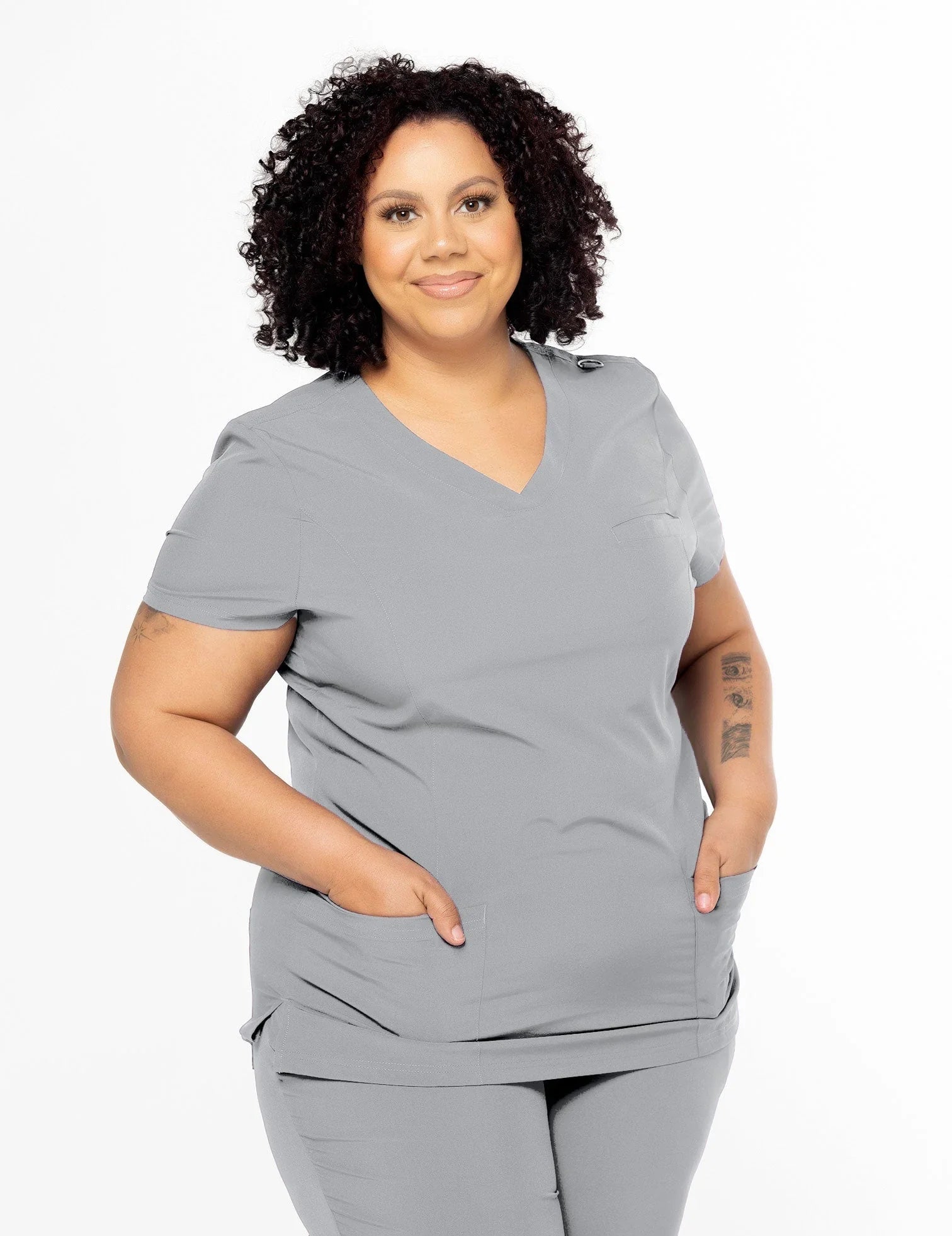 Curly-haired woman wearing gray medical scrubs with pockets, standing smiling against white background