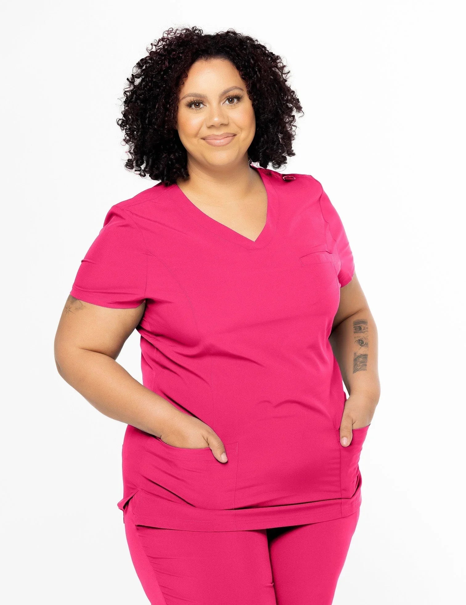 Curly-haired woman wearing hot pink medical scrubs with short sleeves, standing against white background