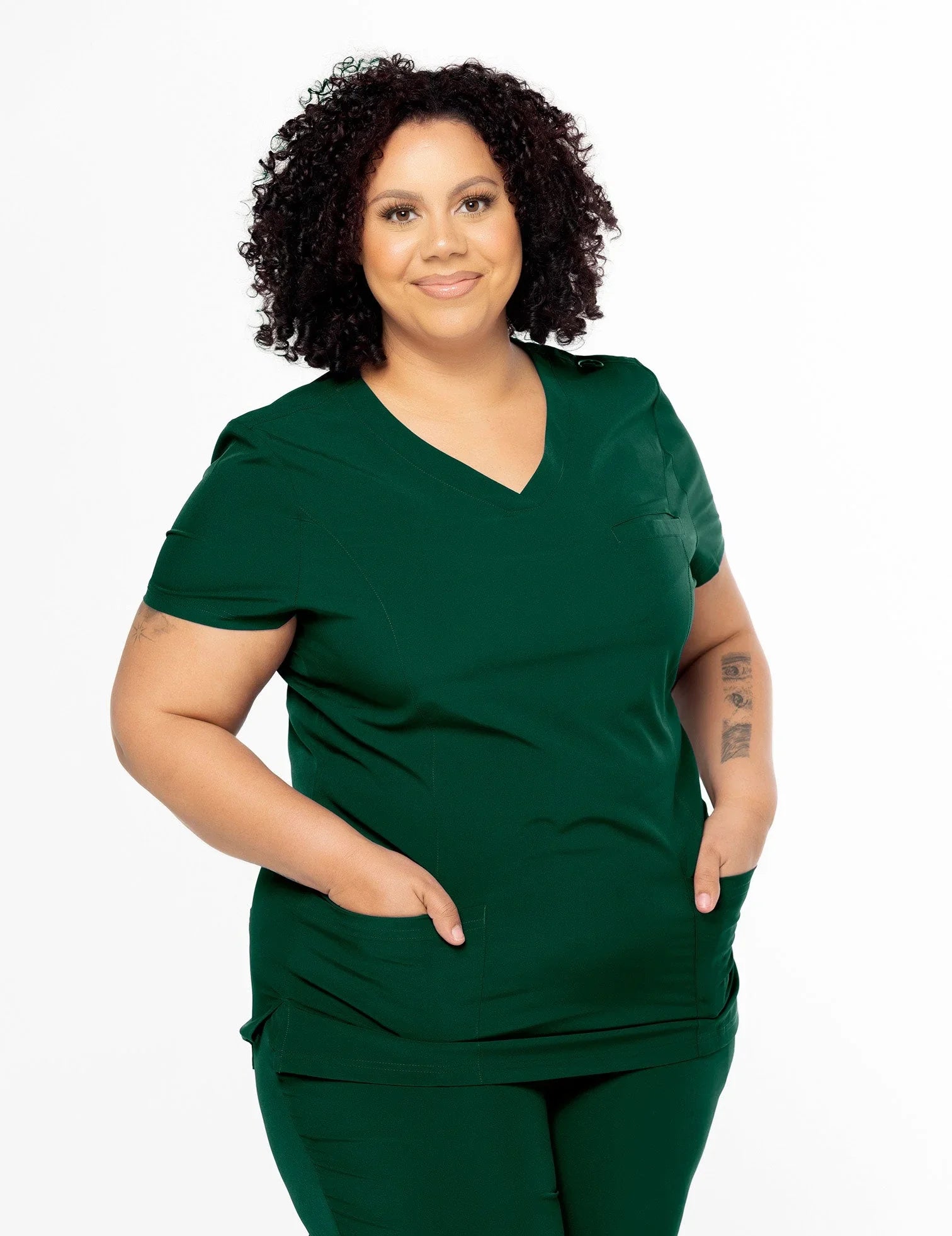 Smiling woman with curly hair wearing dark green medical scrubs with pockets on white background