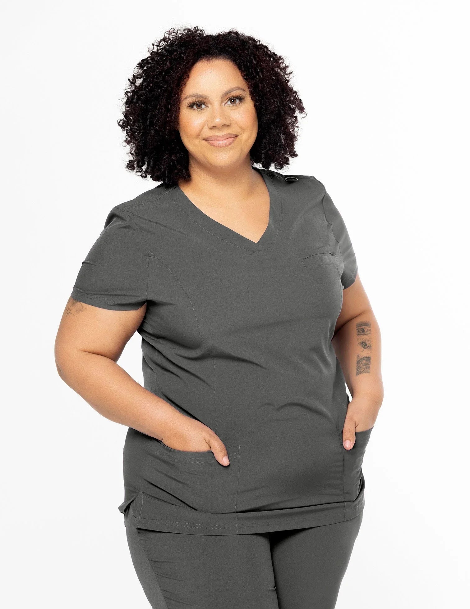 Curly-haired woman wearing pewter-colored medical scrubs with V-neck top and pants, hands in pockets