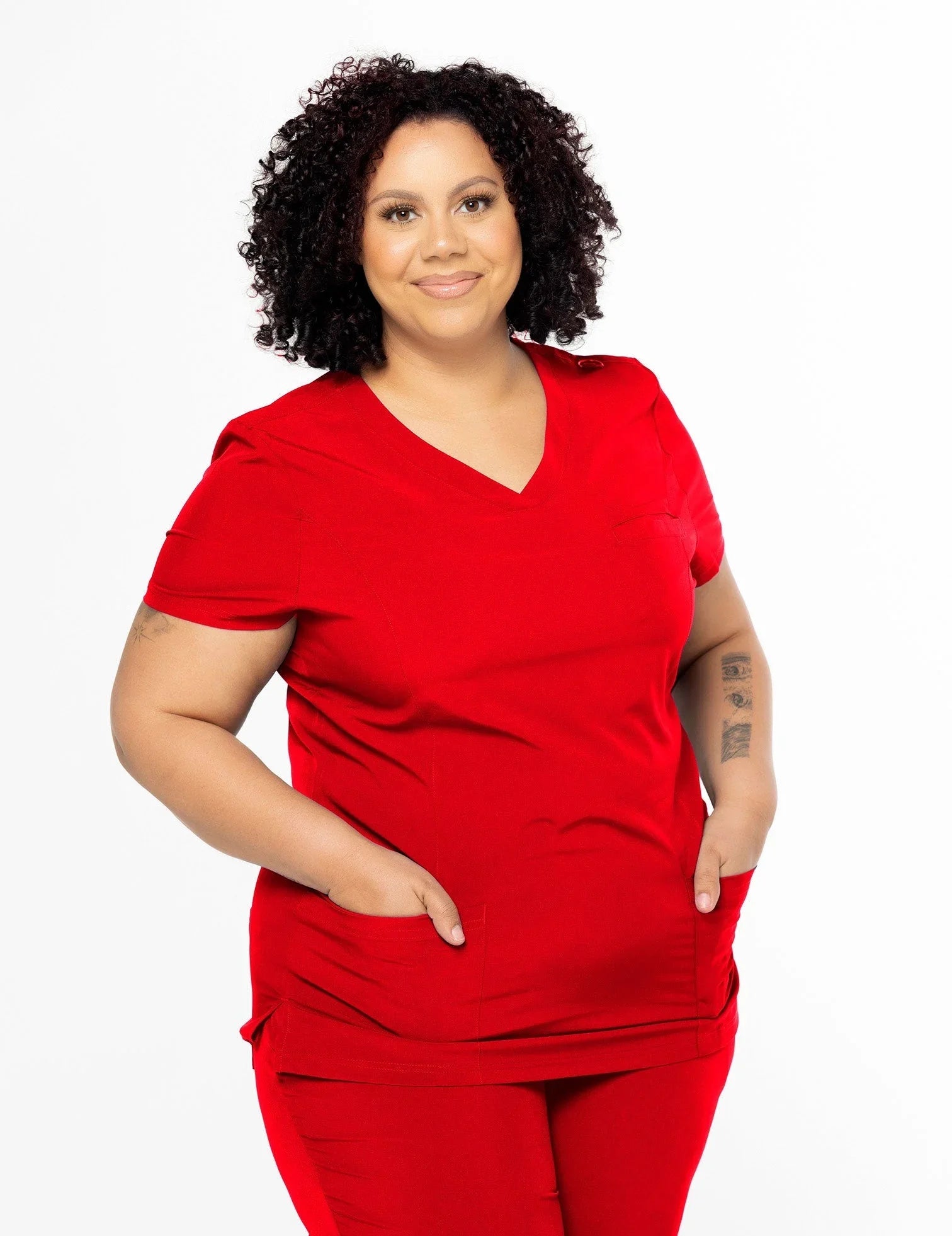 Curly-haired woman wearing red medical scrubs with pockets, smiling against white background